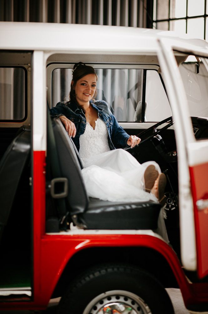 A bride in a white dress and denim jacket sitting inside a classic red and white VW camper van.