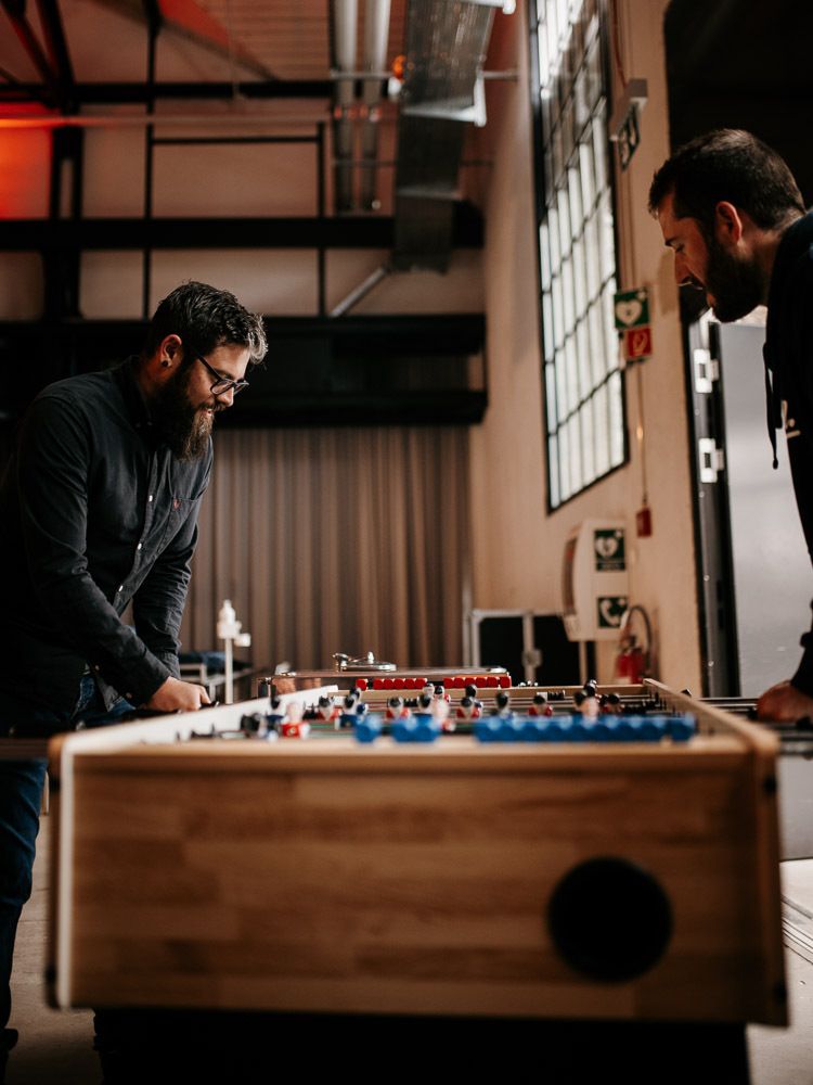 Two bearded men facing each other playing foosball in a modern industrial office space.