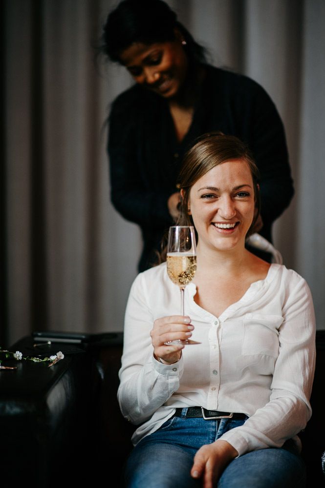 Smiling bride holding a champagne glass while getting her hair styled before the wedding.