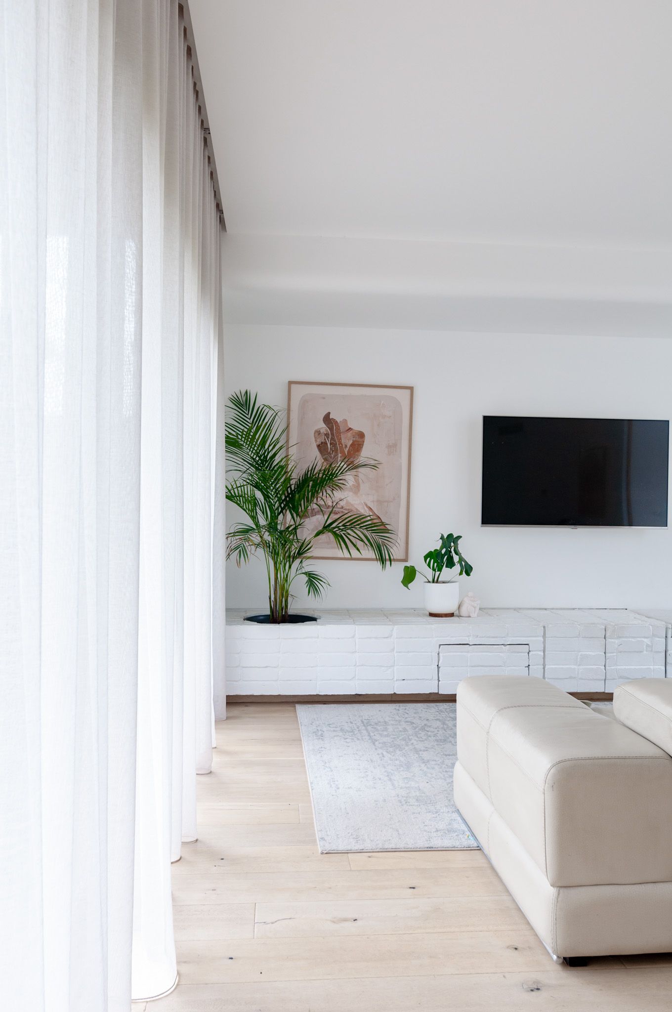 Contemporary living room with wall-mounted TV, indoor plants, and white furniture against light wood floors.
