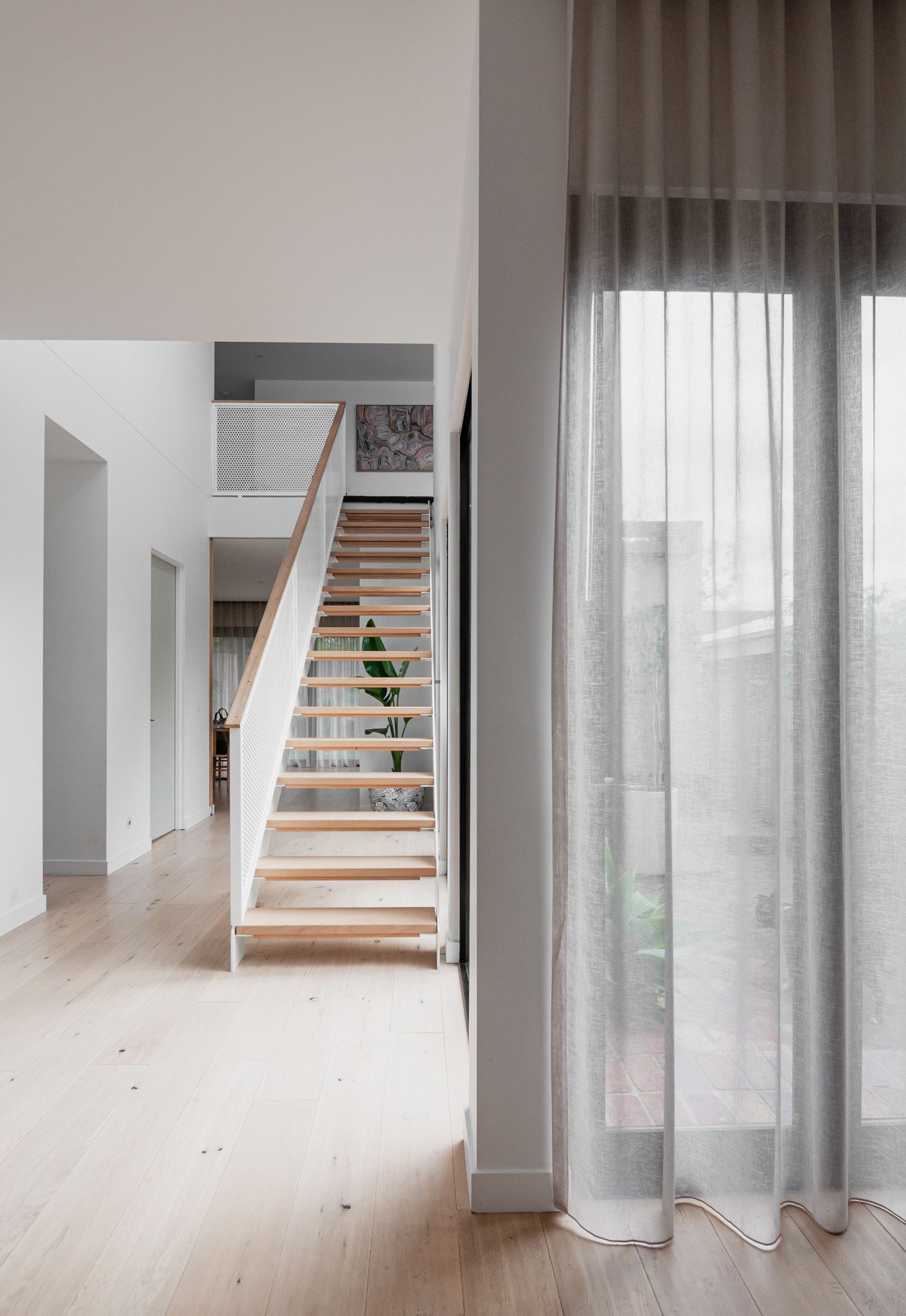 Minimalist hallway featuring wooden staircase with white railings and sheer grey curtains.