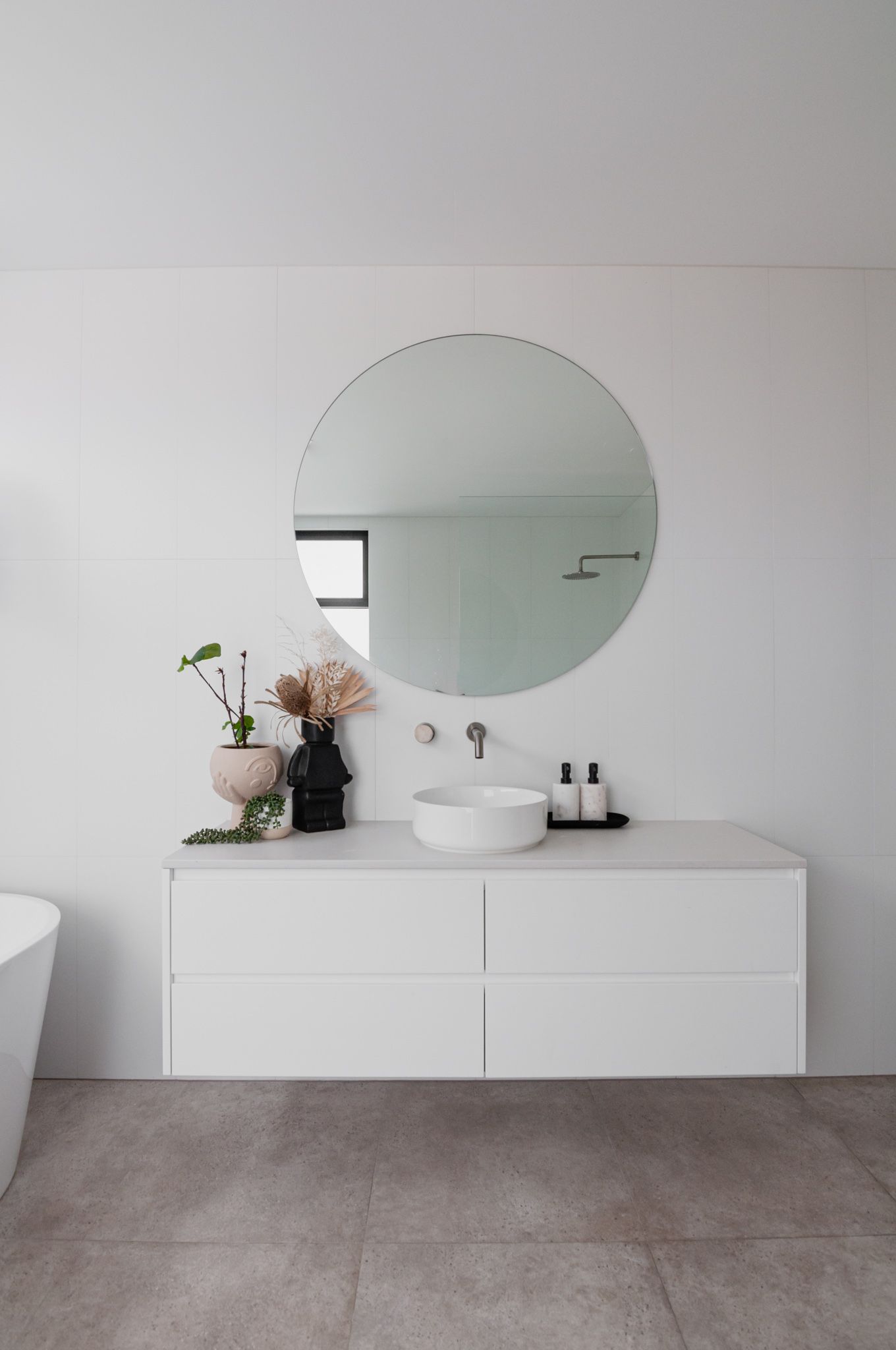 Modern minimalist bathroom vanity with round mirror, white drawers, and floral accents on a grey floor.