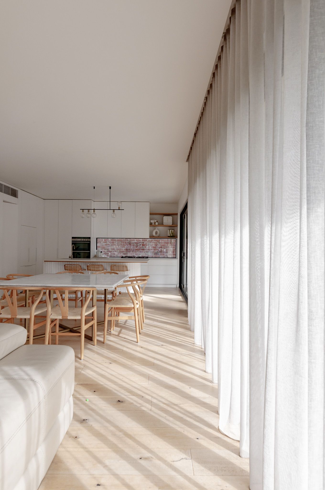 Bright open concept dining area with wooden chairs and floor-to-ceiling white curtains.
