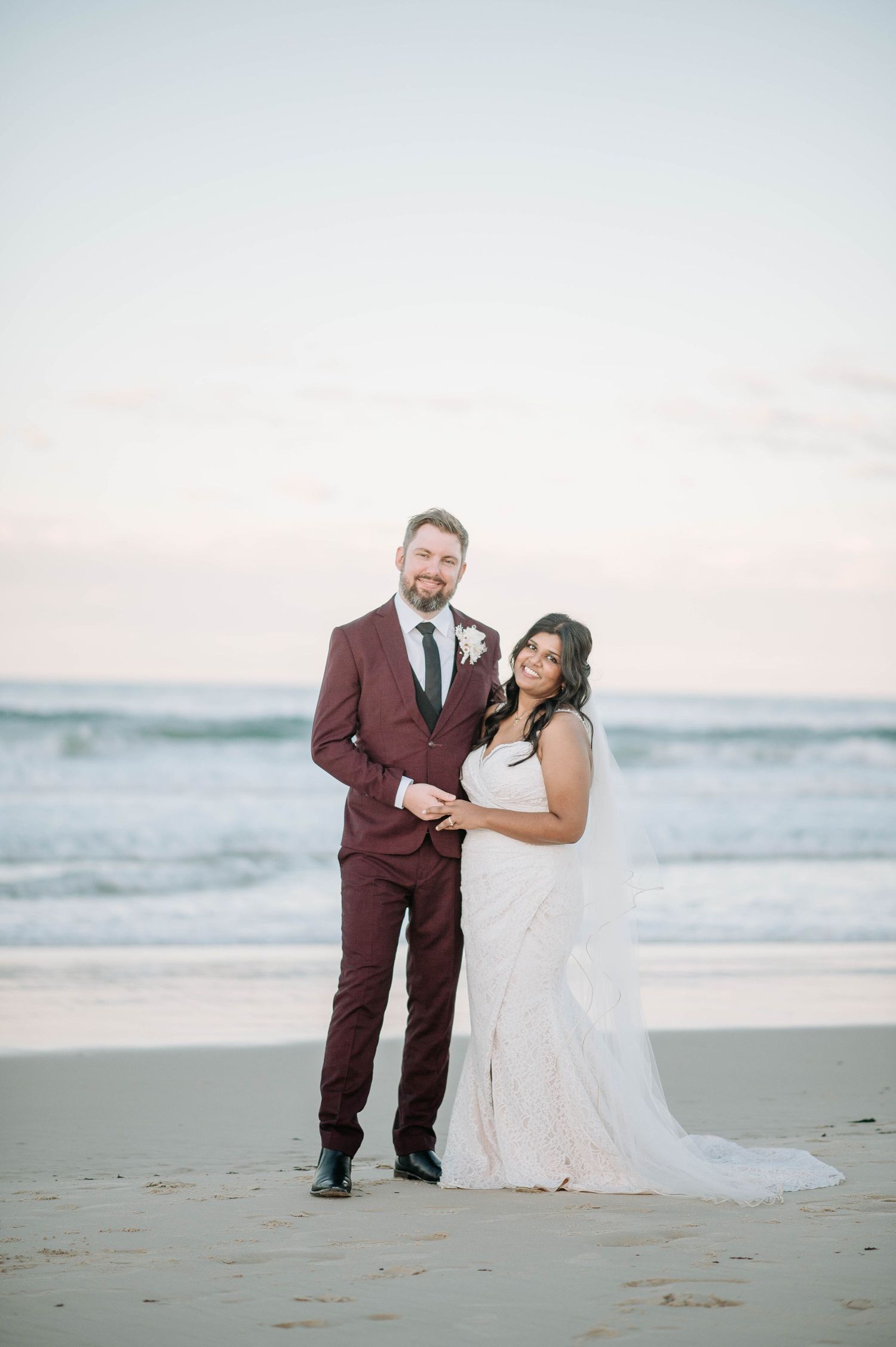 Elegant beach wedding portrait with couple in burgundy suit and white gown.