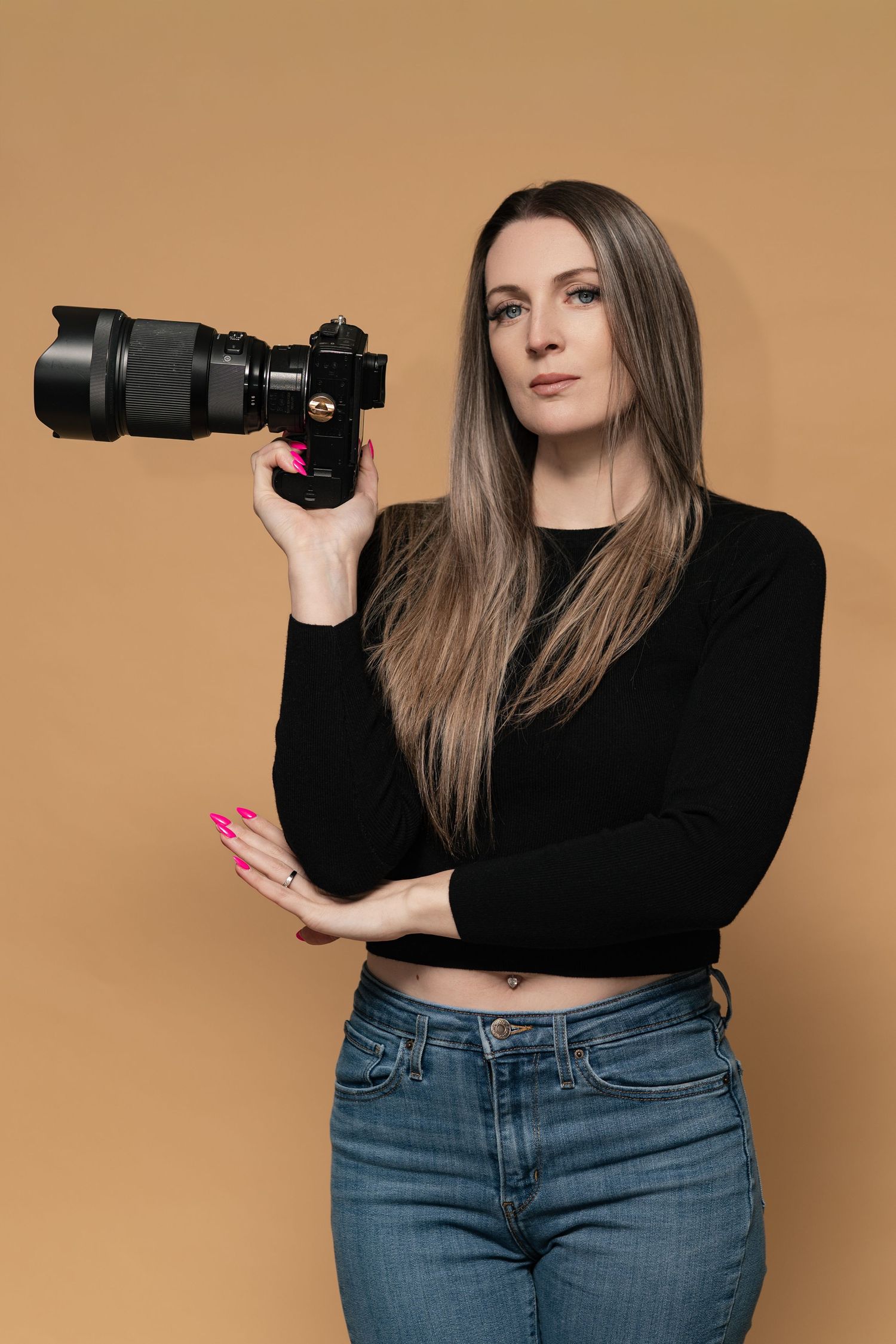 Professional photographer in black top and jeans poses with camera against beige backdrop in studio sequence.