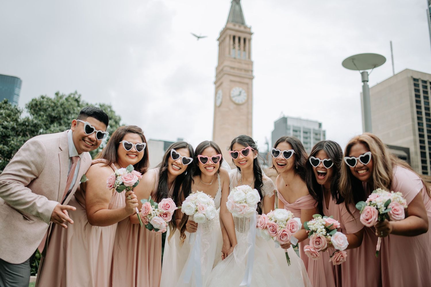 Wedding party wearing pink dresses and suits posing together outside with a clock tower in the background.