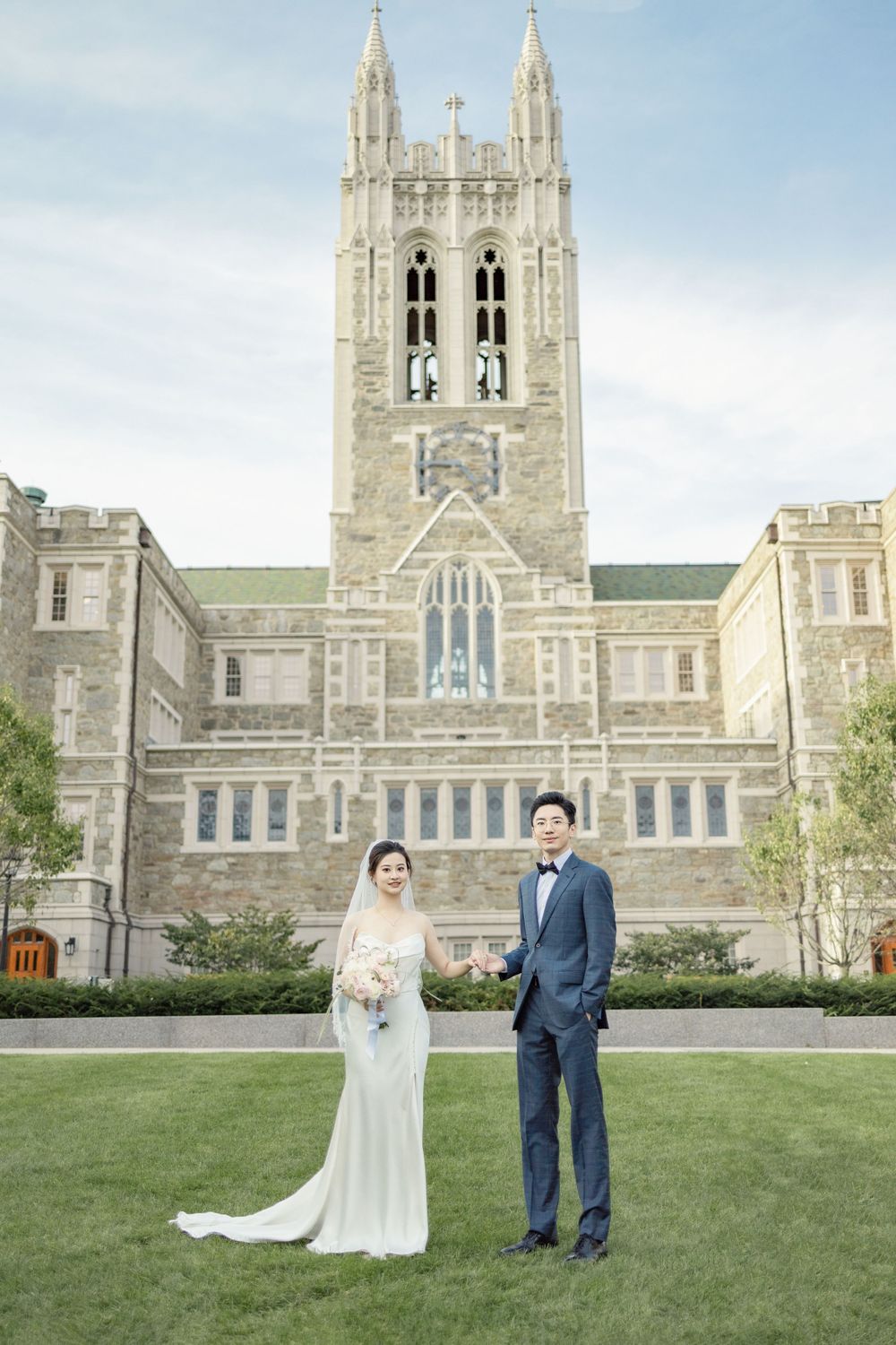 Wedding couple poses on lawn in front of gothic stone university building.
