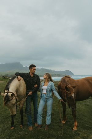 A couple stands with their horses in a grassy field against a cloudy mountain backdrop.