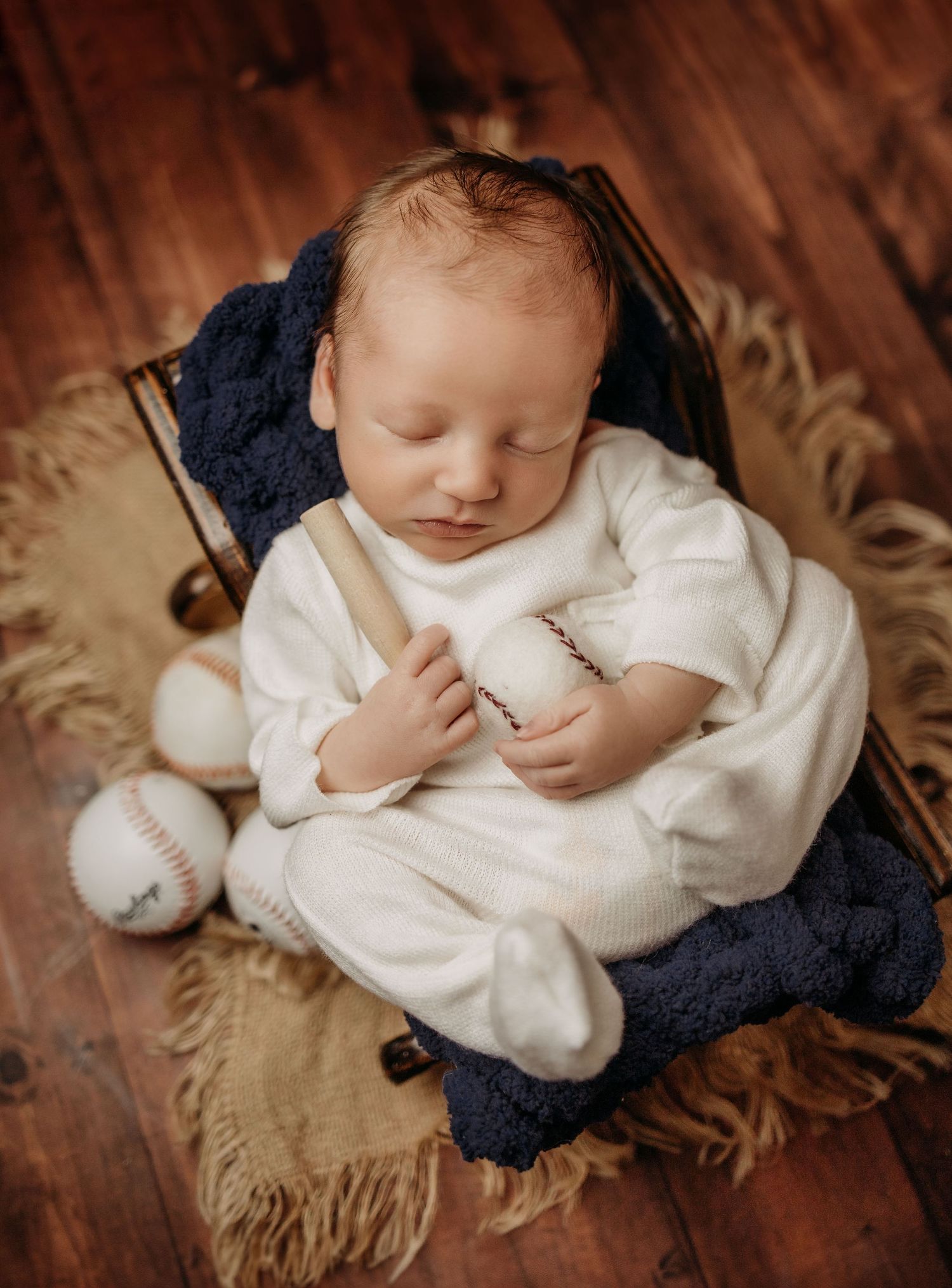 Sleeping newborn with baseball props on furry blanket and wooden floor.