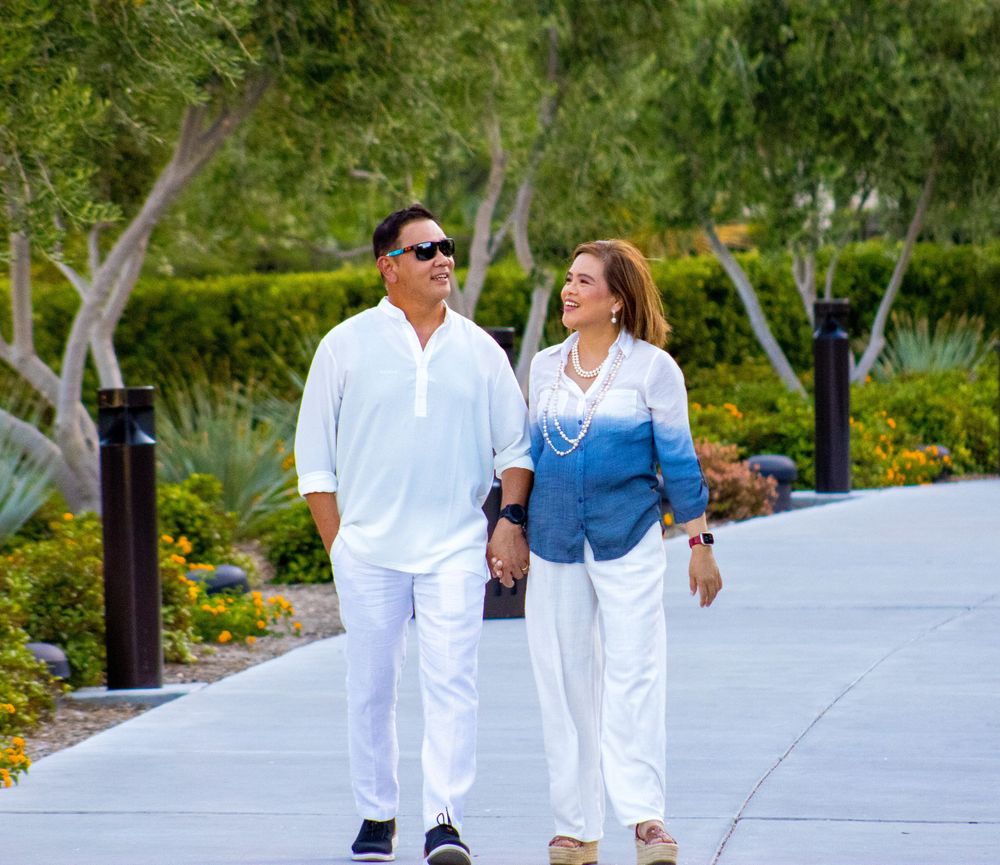 A couple walks hand-in-hand along a sunny garden path, wearing coordinating white and blue outfits.