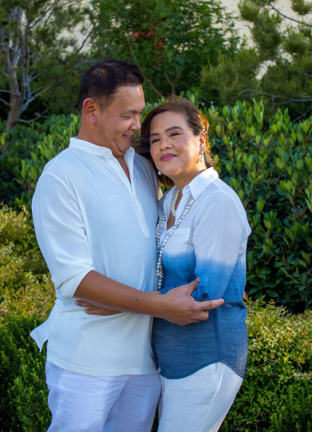 Loving couple in white and blue outfits embracing warmly in a sunlit garden setting.