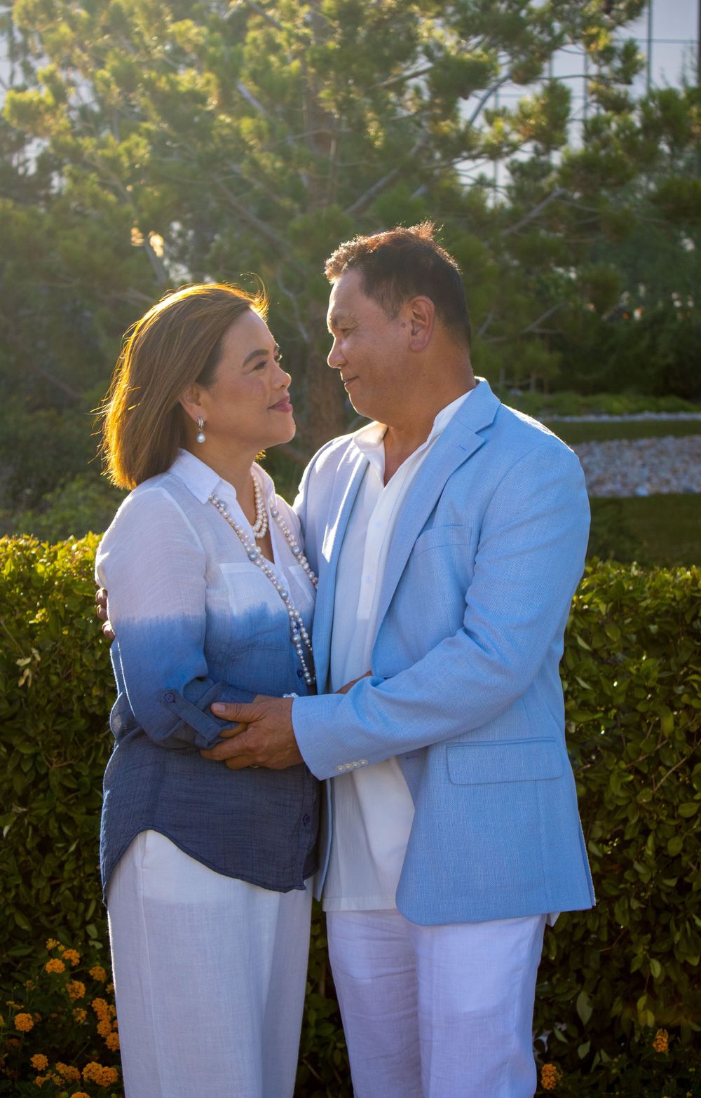 A couple embraces warmly in a sunlit garden, dressed in elegant blue and white outfits.