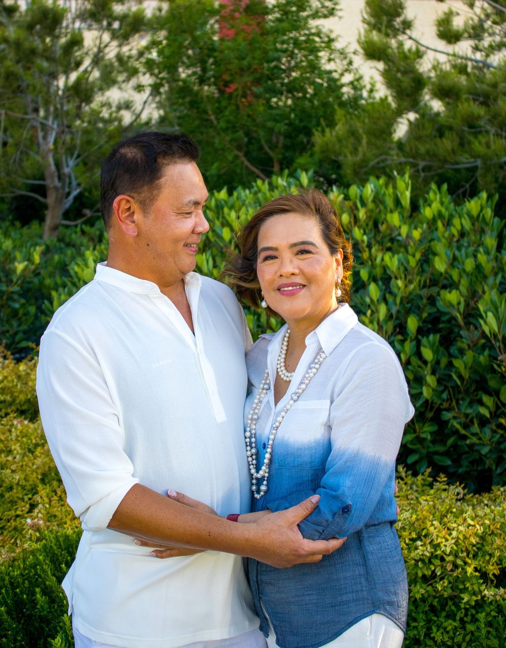 A smiling couple poses together outdoors, the woman wearing a blue ombre shirt with pearl necklace.