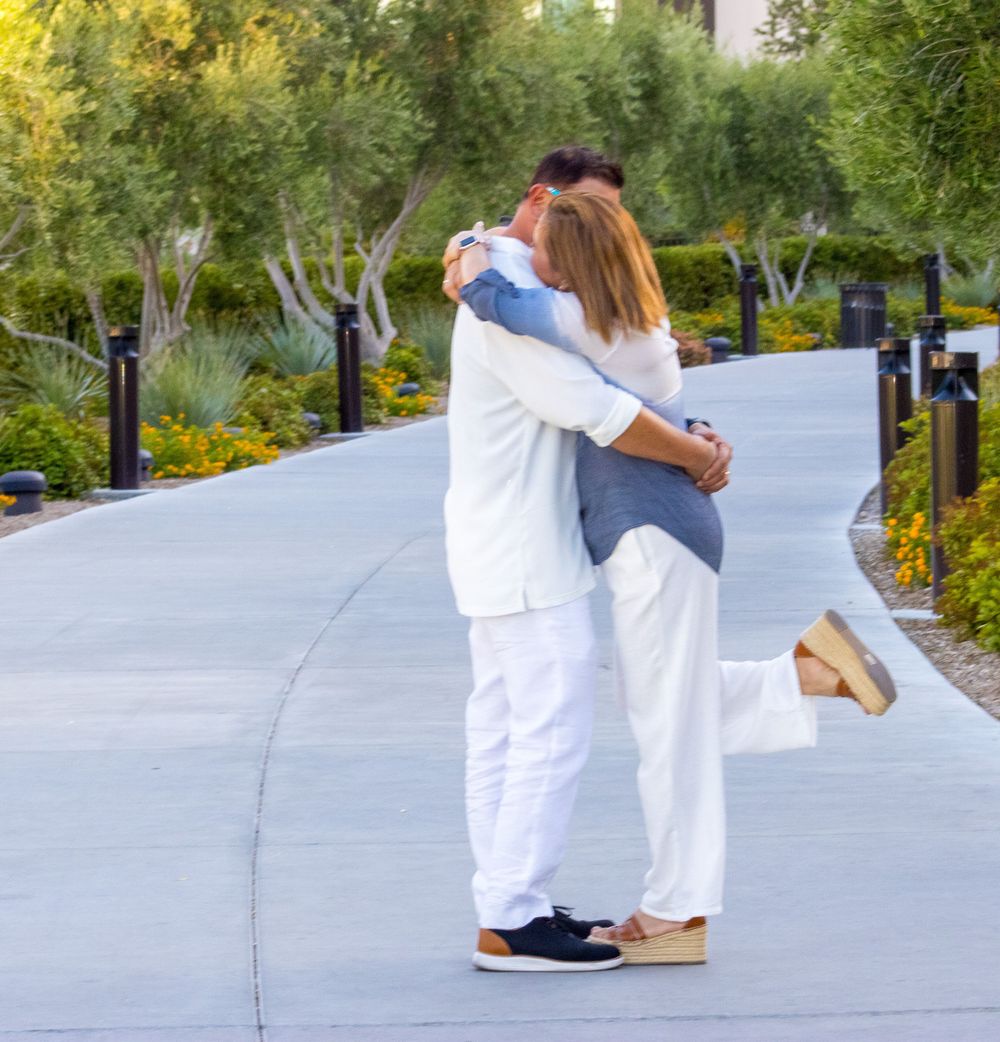 A couple shares a romantic embrace and kiss on a winding garden pathway lined with olive trees.