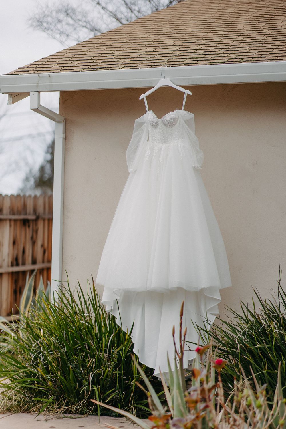 White wedding dress hanging on exterior wall with garden foliage below.