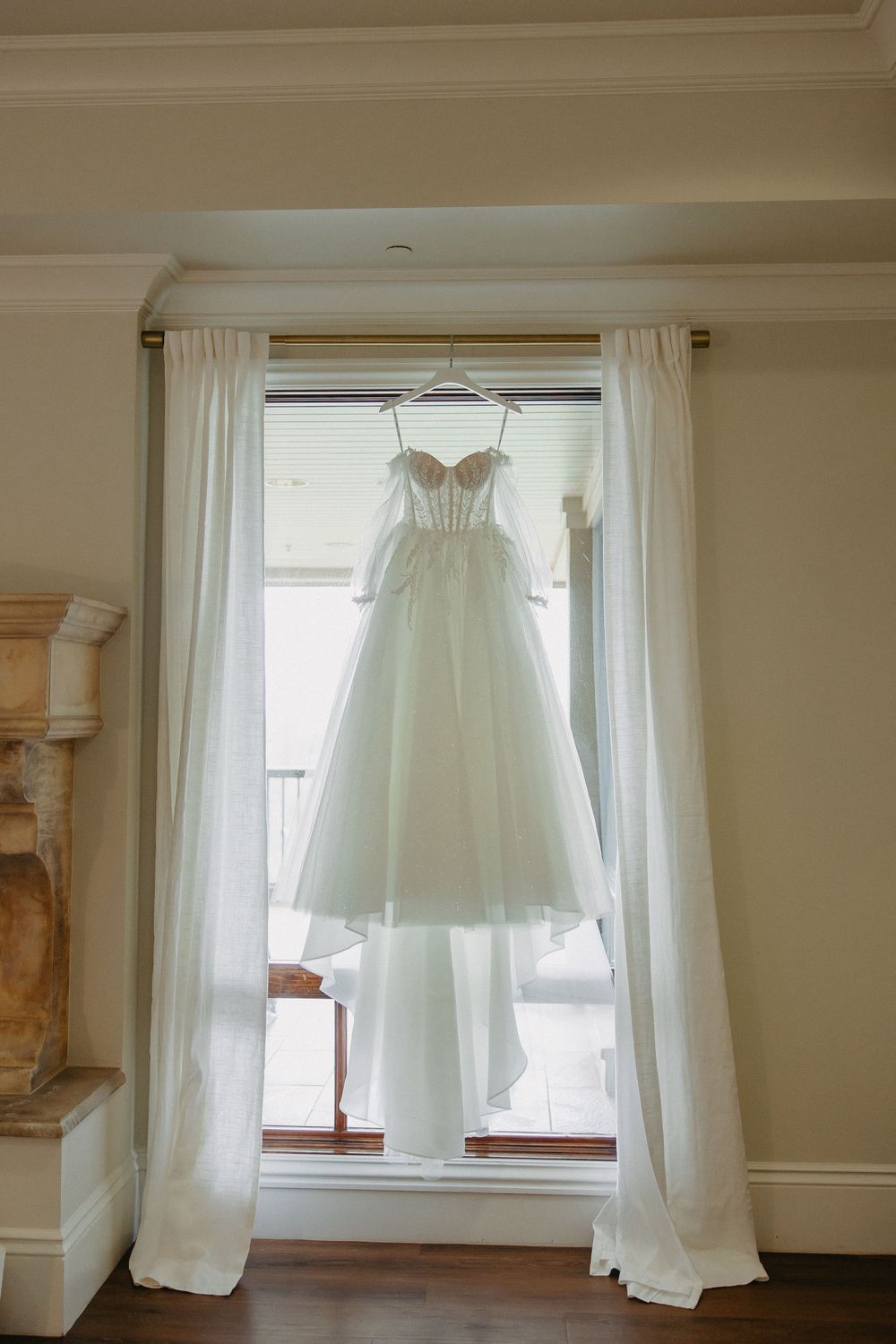White wedding gown hanging in front of window with sheer curtains in elegant room setting.