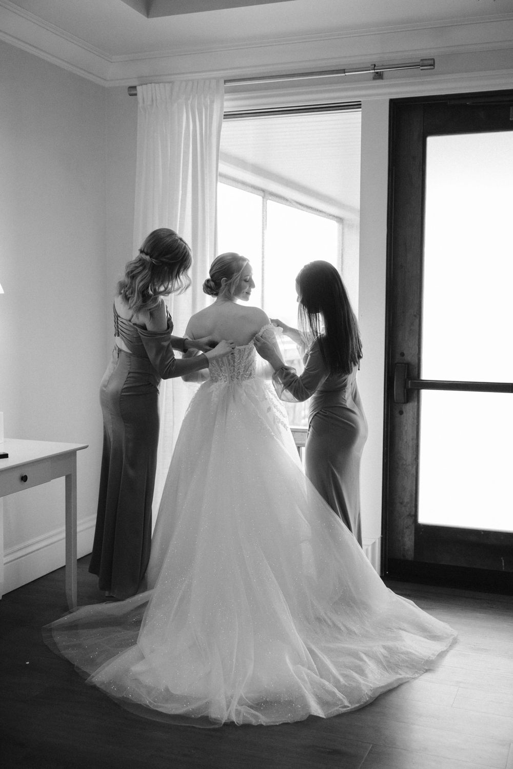 Black and white photo of wedding dress preparation with attendants helping adjust the gown.