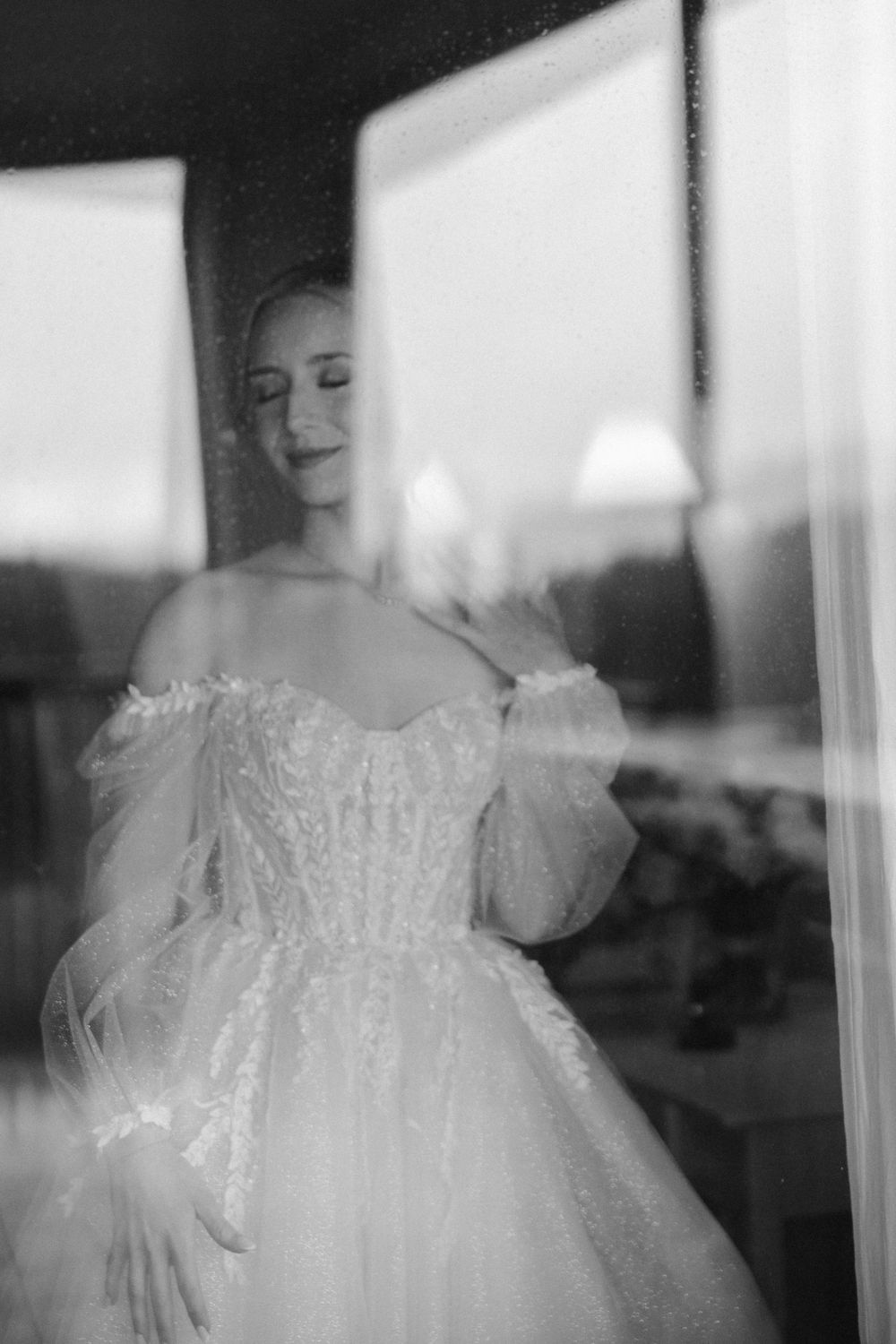 Artistic black and white portrait through window reflection showing bride in wedding dress.
