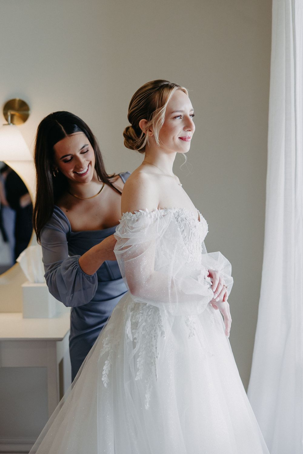 Elegant wedding dress preparation moment with bridesmaid helping adjust the gown.