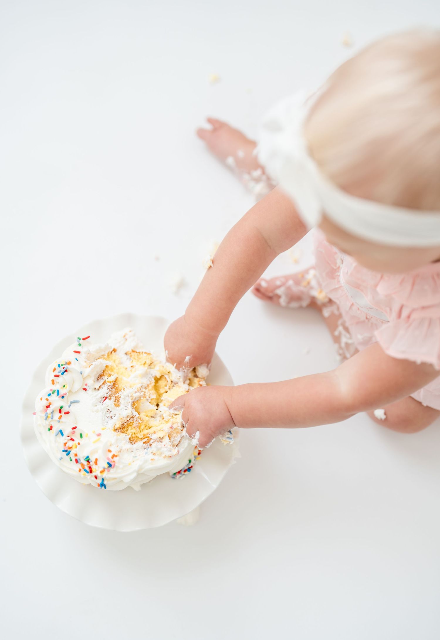 Smiling toddler in white dress resting chin on hands while sitting at table.