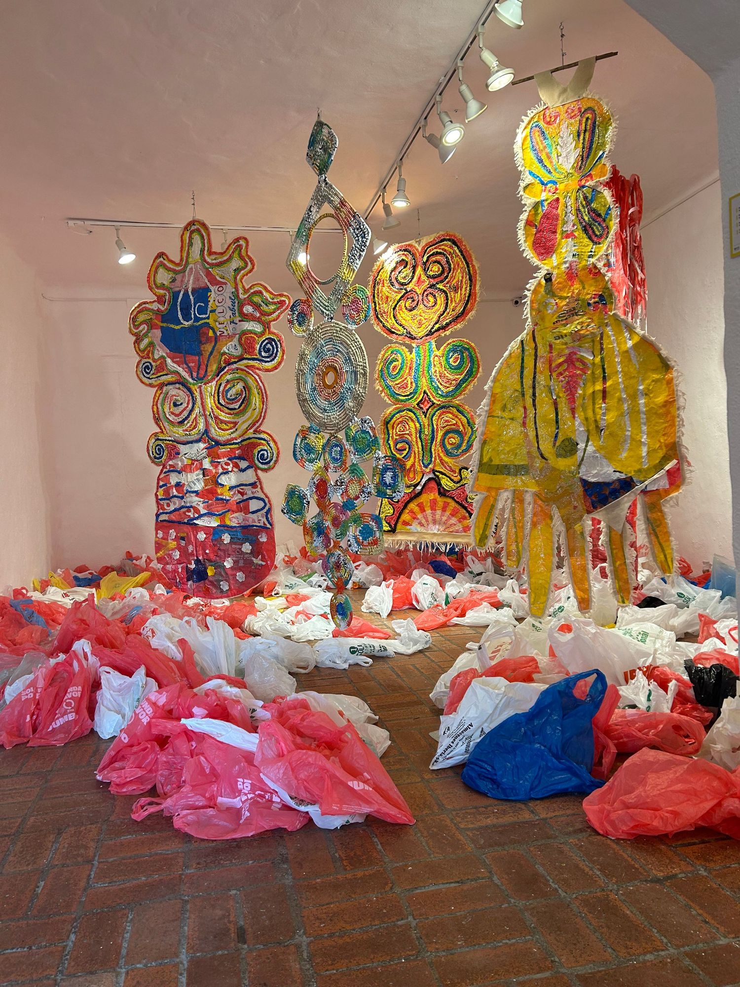 Colorful gallery installation with large decorative sculptures and scattered red and blue plastic bags on brick floor.