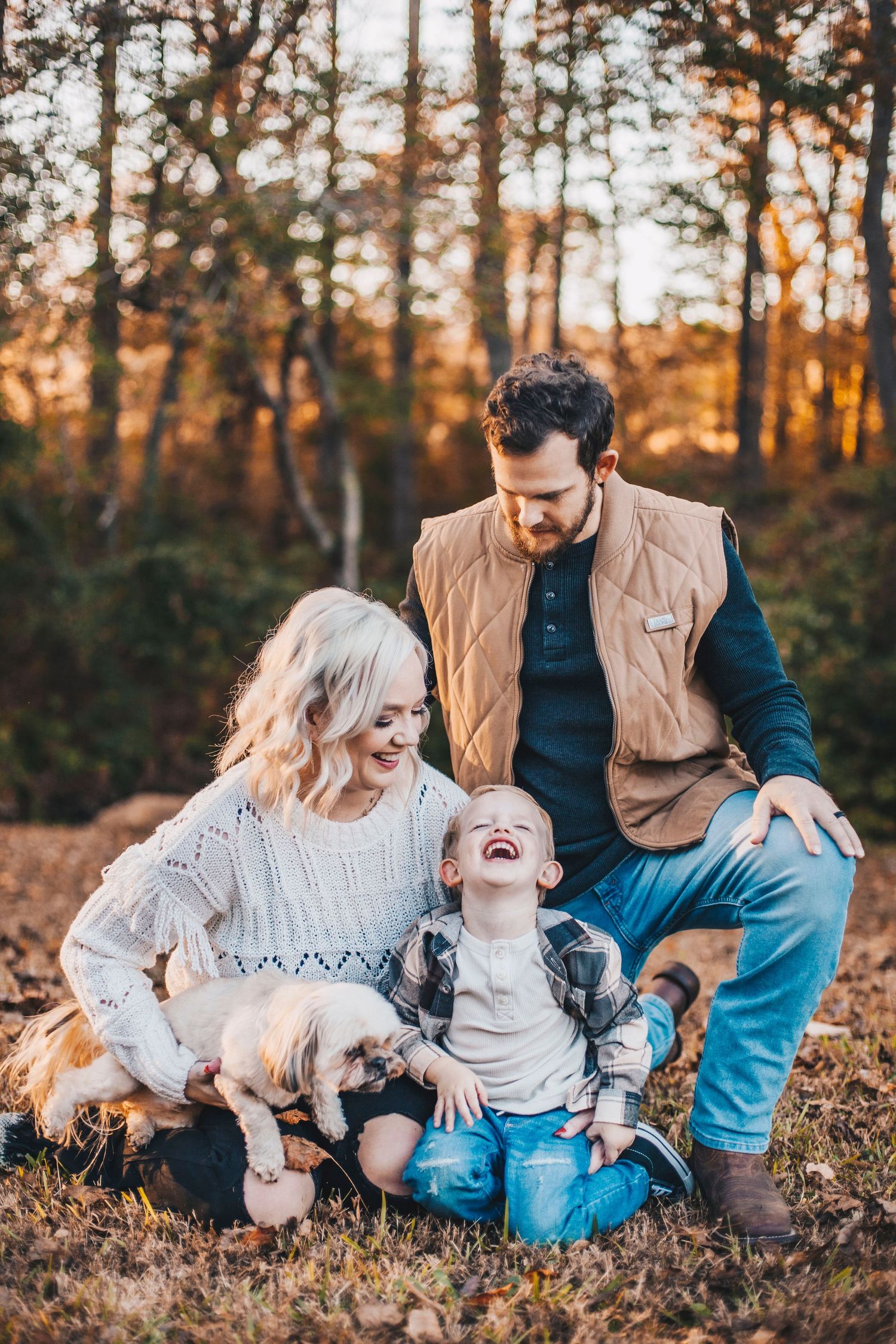 Happy family with young child laughing together in autumn forest at golden hour.
