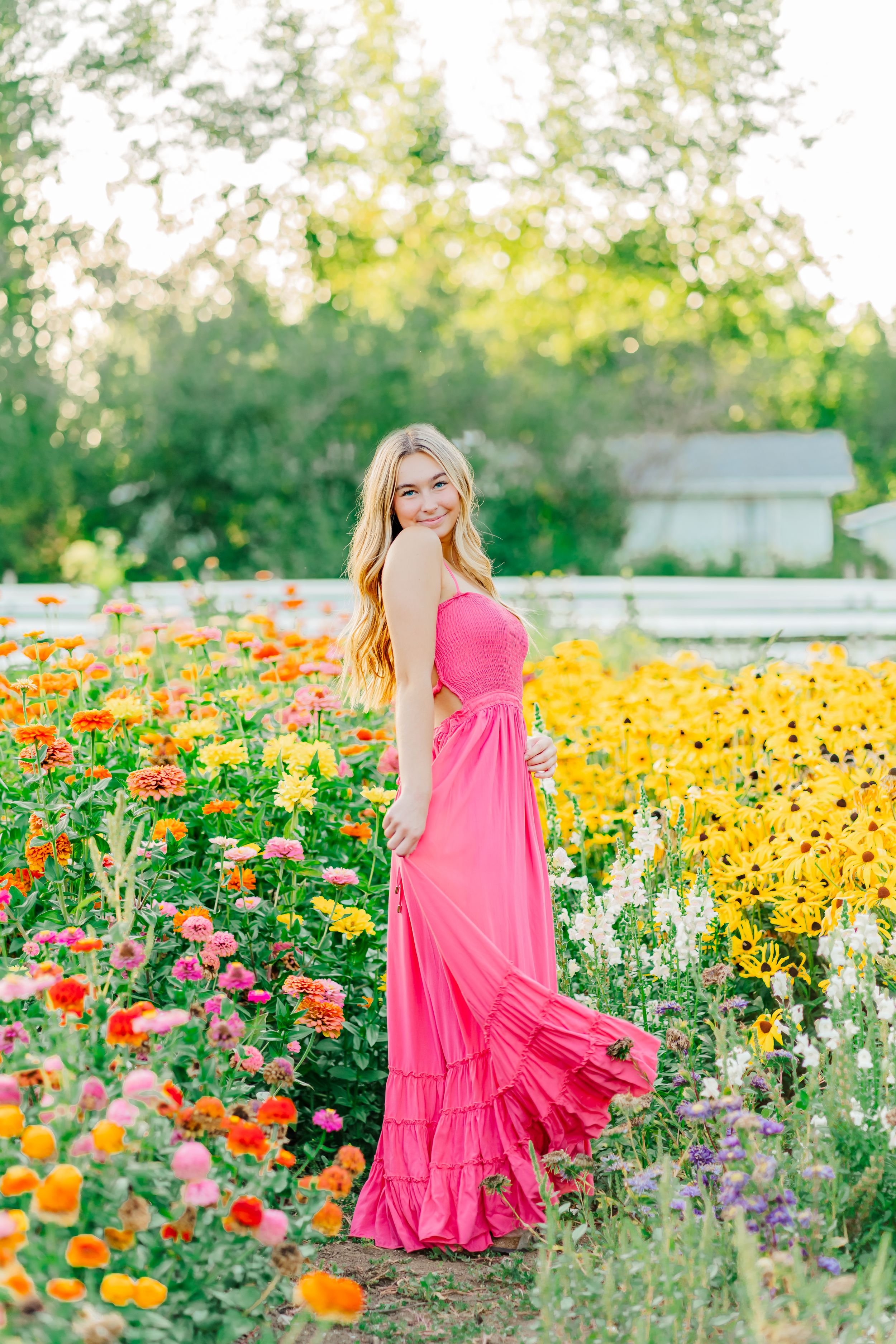 A vibrant summer garden blooms with orange and yellow flowers surrounding a senior girl in a flowing pink maxi dress in natural light.