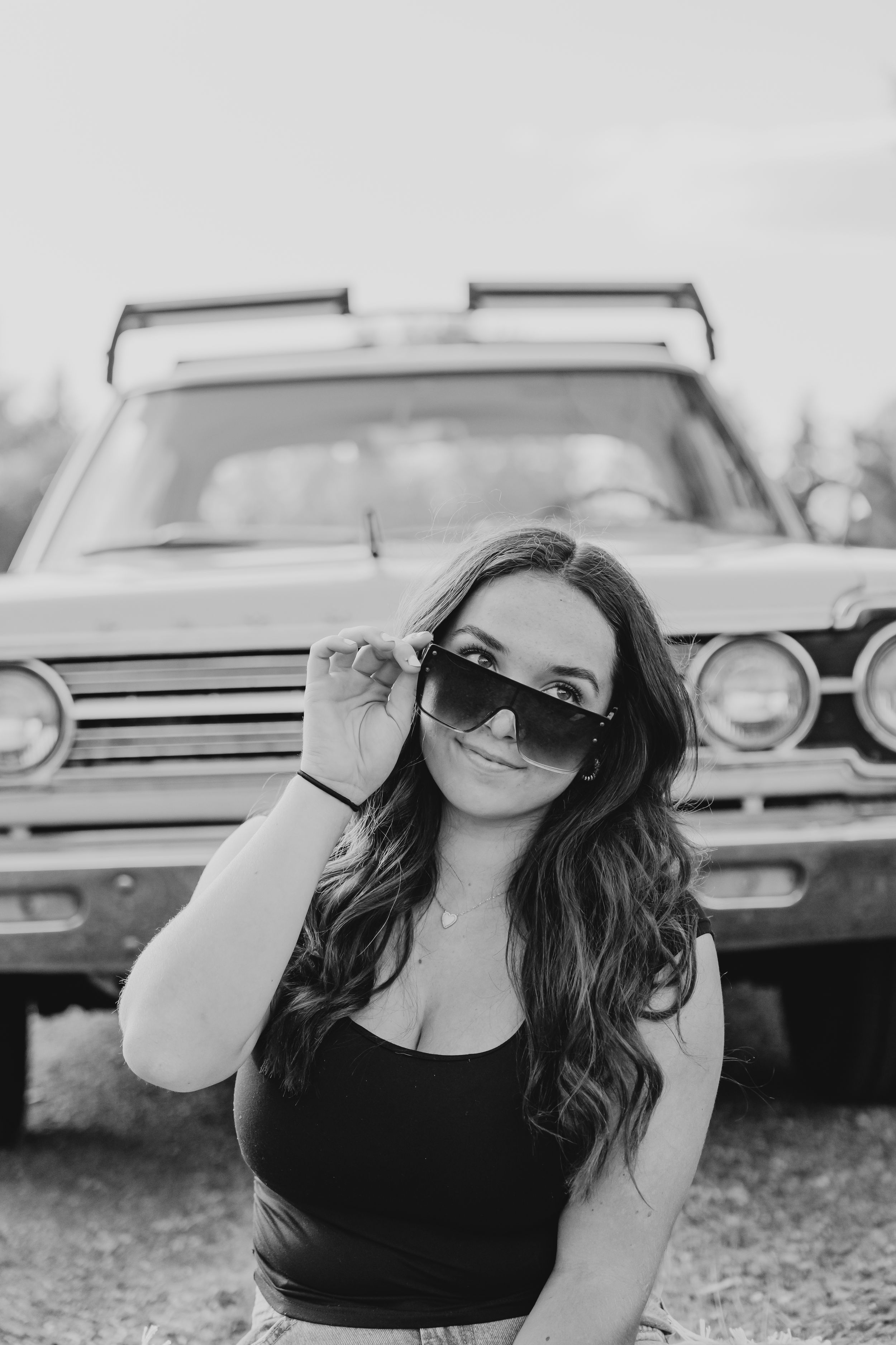 Black and white portrait of senior girl adjusting sunglasses while posing in front of a vintage car.