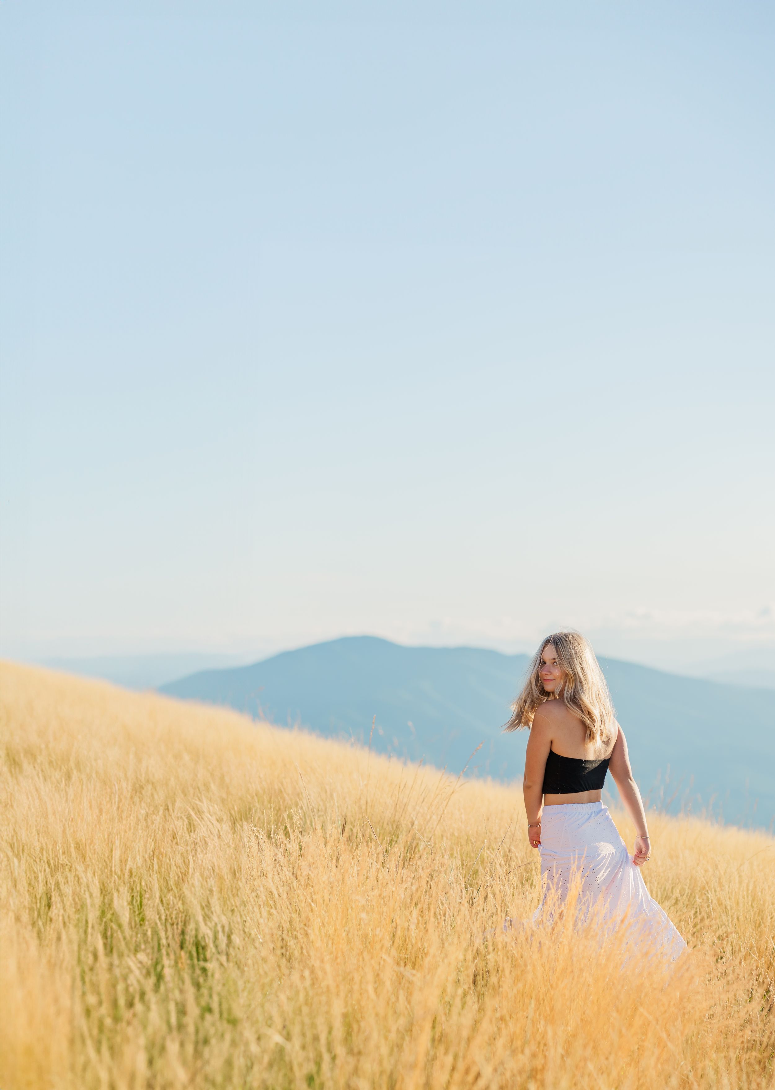 High School senior girl in a white skirt stands among golden grass overlooking distant mountains on a sunny day.