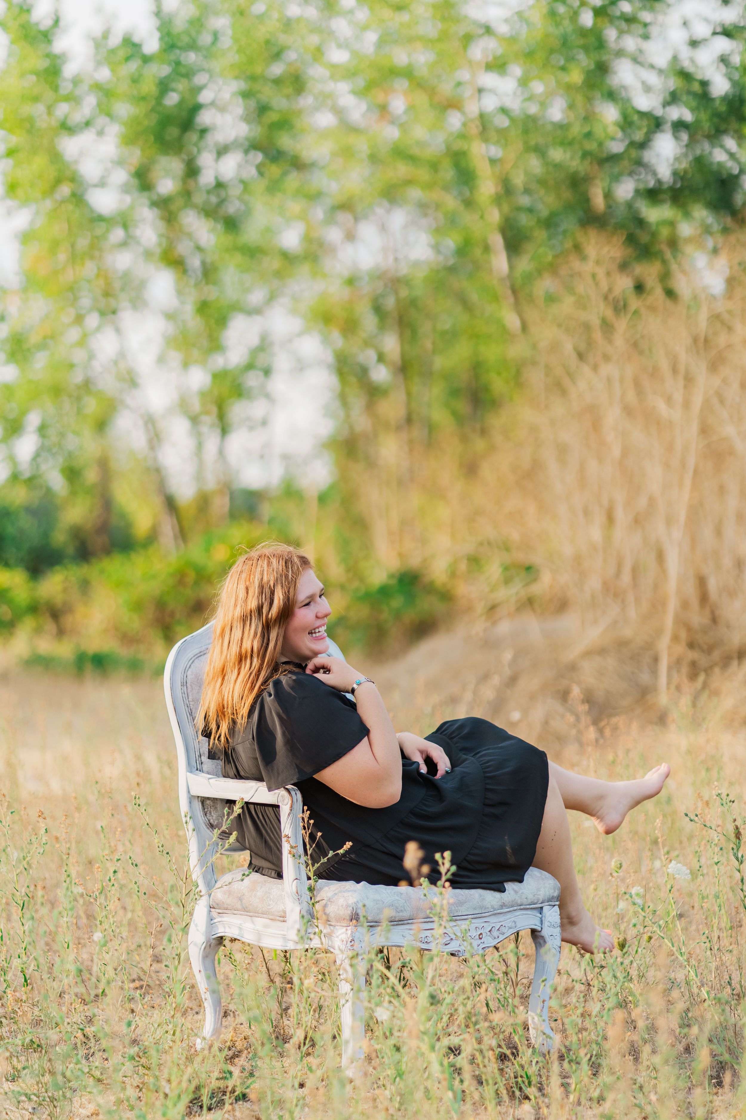 A senior girl sits playfully in vintage white chair in a meadow with tall grass and trees, creating a dreamy outdoor scene.