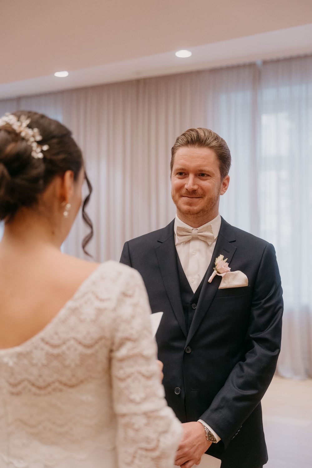 A groom in a black tuxedo and bow tie shares a joyful first look moment with his bride in a lace wedding gown.