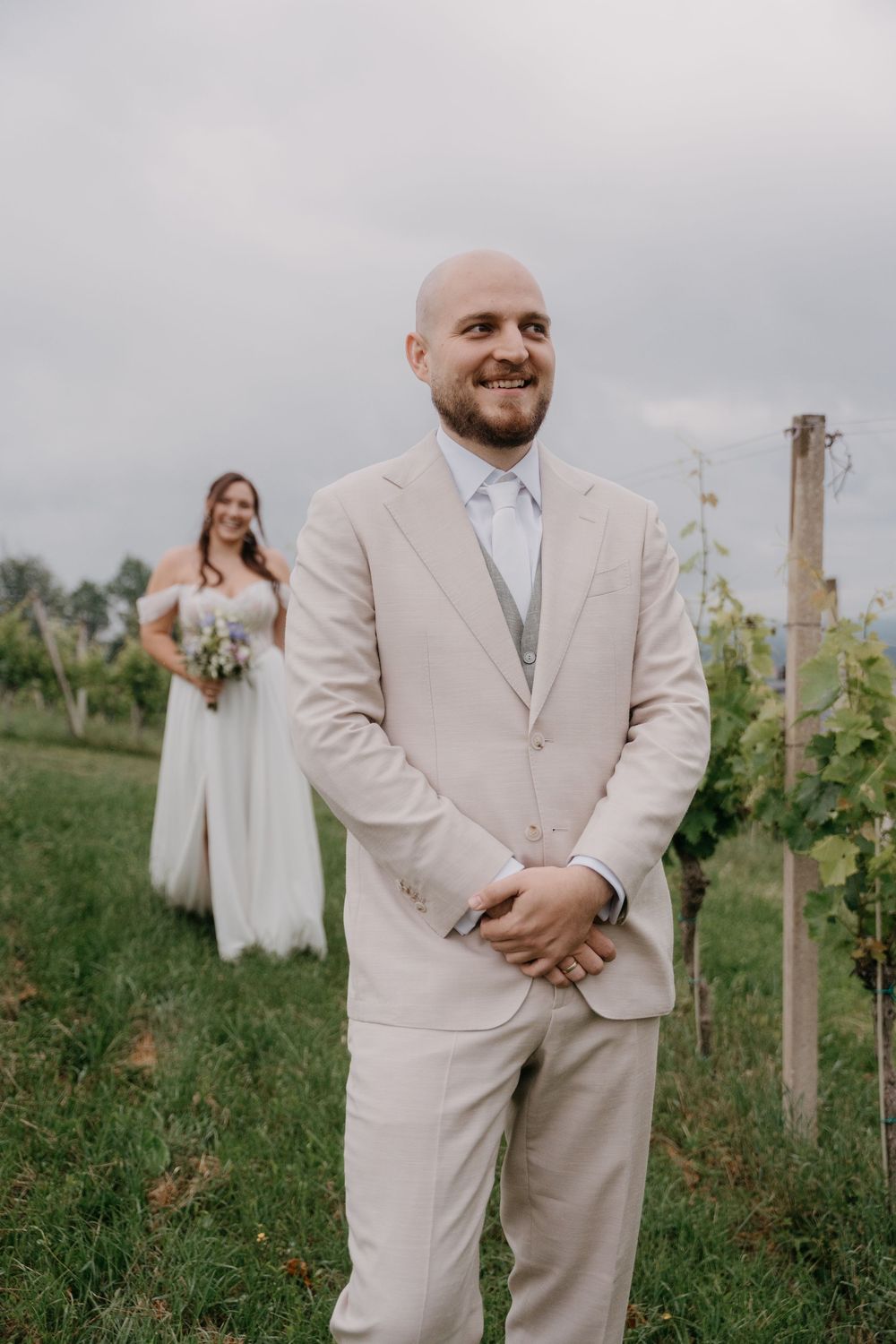 Groom in beige suit smiling at vineyard wedding, bride in white dress holding bouquet standing in background.