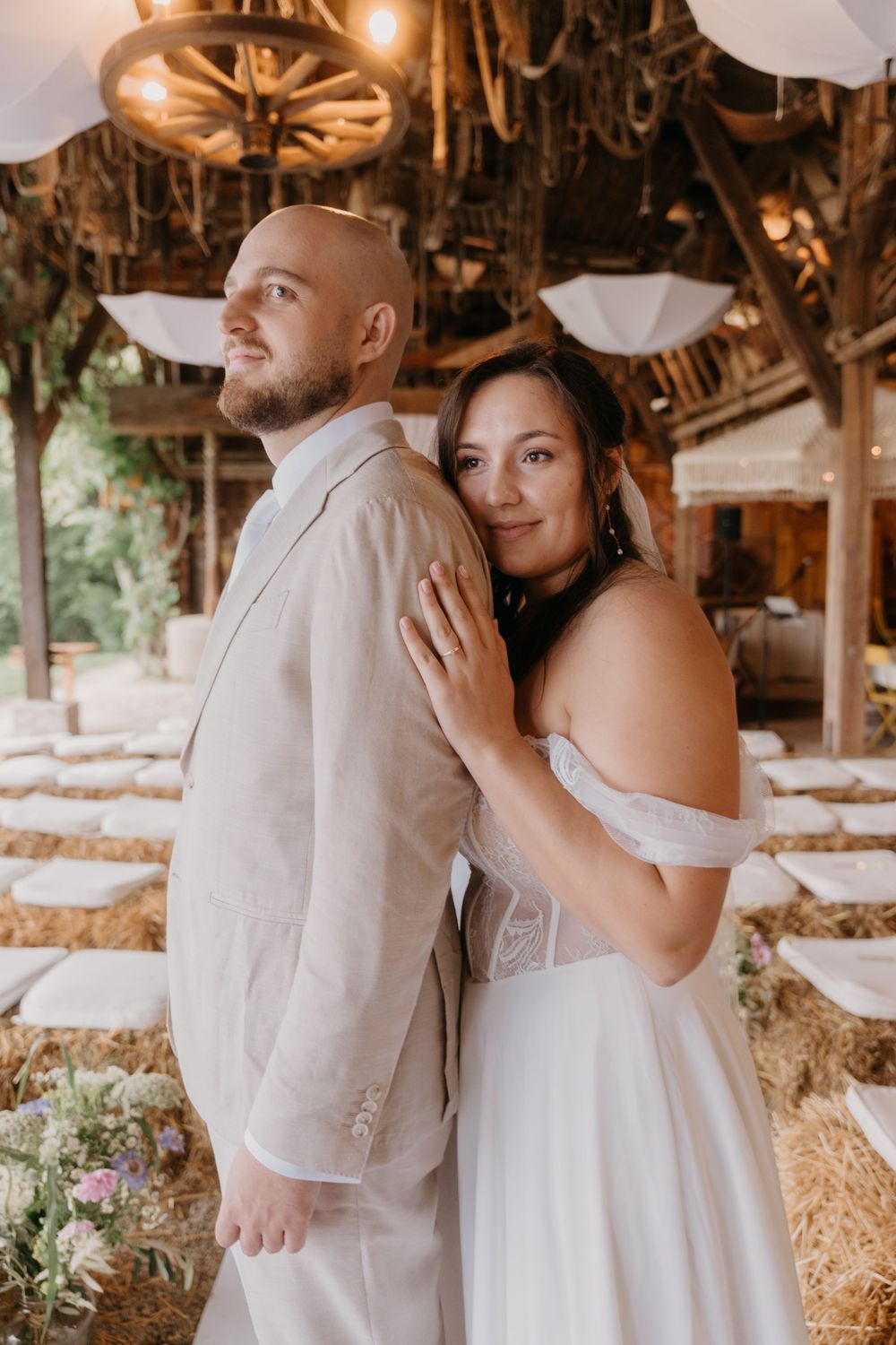 Bride and groom pose back-to-back in a rustic barn wedding venue with hay bales and wagon wheel chandeliers.