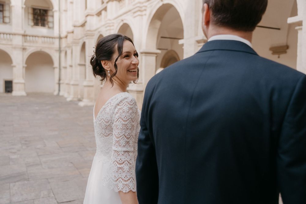 Bride and groom share an intimate moment in a courtyard with stone arches and classical architecture.