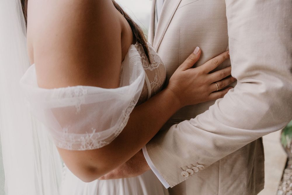 Close-up of bride and groom embracing, showcasing lace wedding dress and beige suit details.
