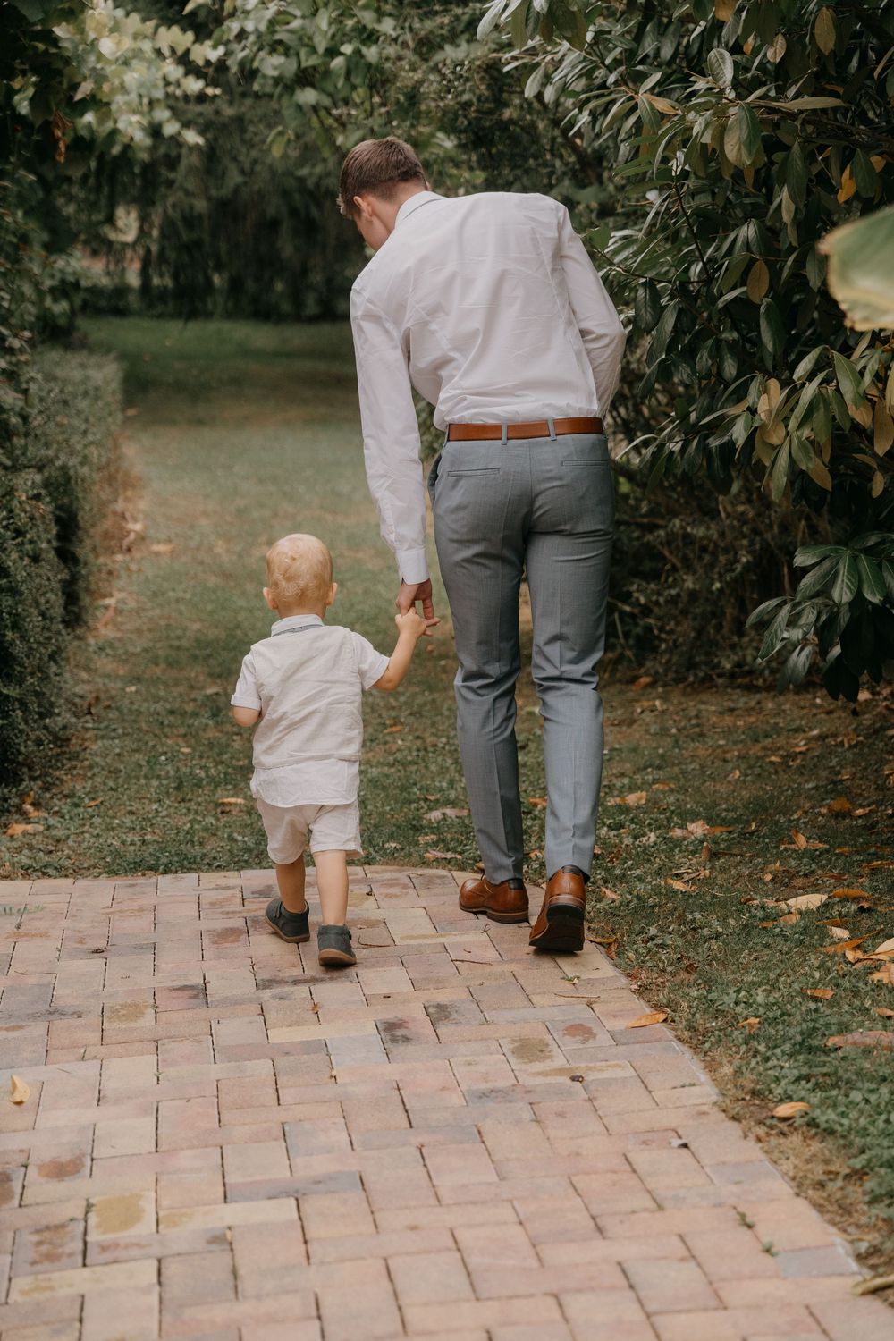 Parent and toddler walking together on a brick pathway through a garden setting, viewed from behind.