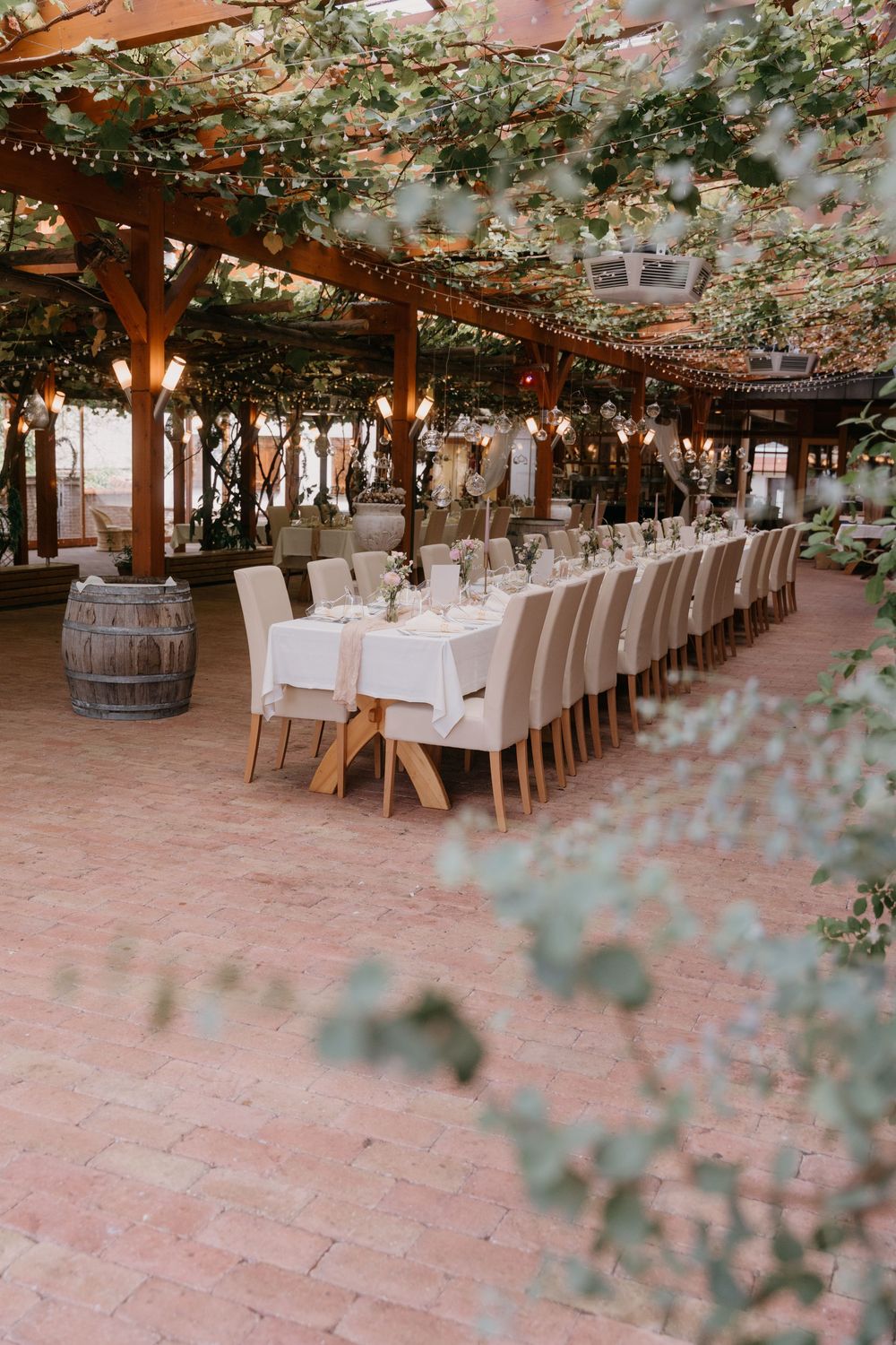 Long dining tables set for an elegant outdoor wedding reception under a rustic wooden pergola with floral decorations.