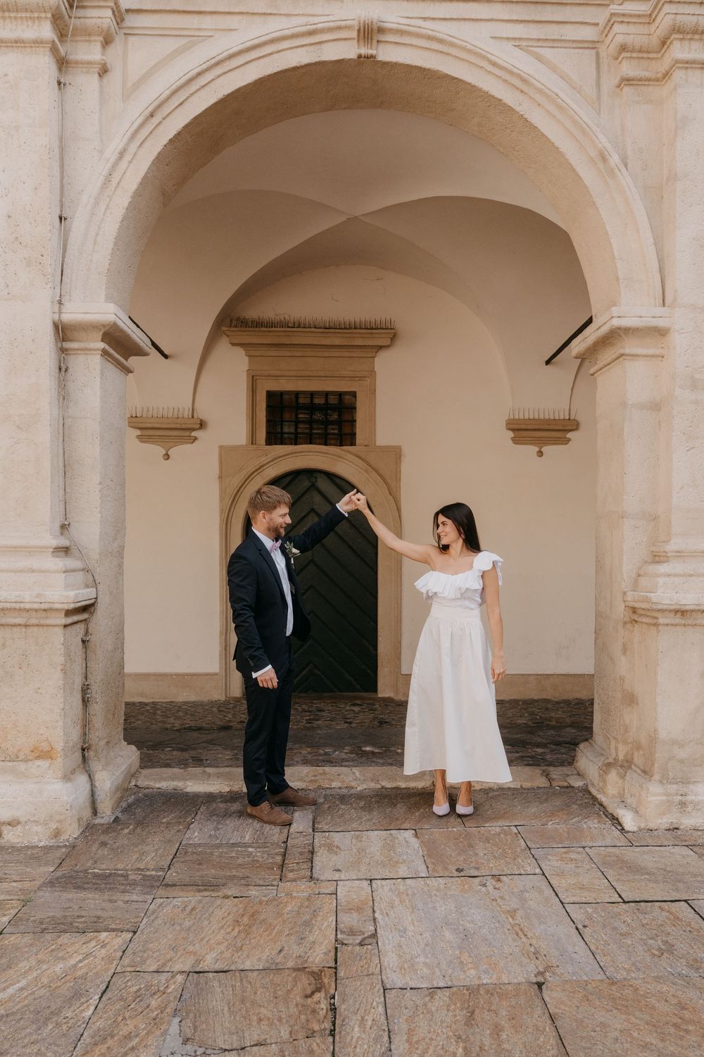A couple shares a playful moment under white stone archways in a historic architectural setting.