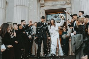 Wedding party celebrates outside historic columned building as guests throw confetti in joyful black and white moment.