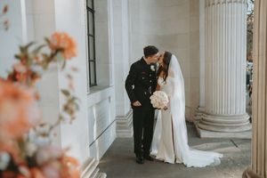 A romantic wedding couple shares intimate moments in an elegant white columned venue with soft peach floral accents.
