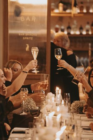 A candlelit dinner table setting with people raising wine glasses in celebration at a dimly lit restaurant bar.
