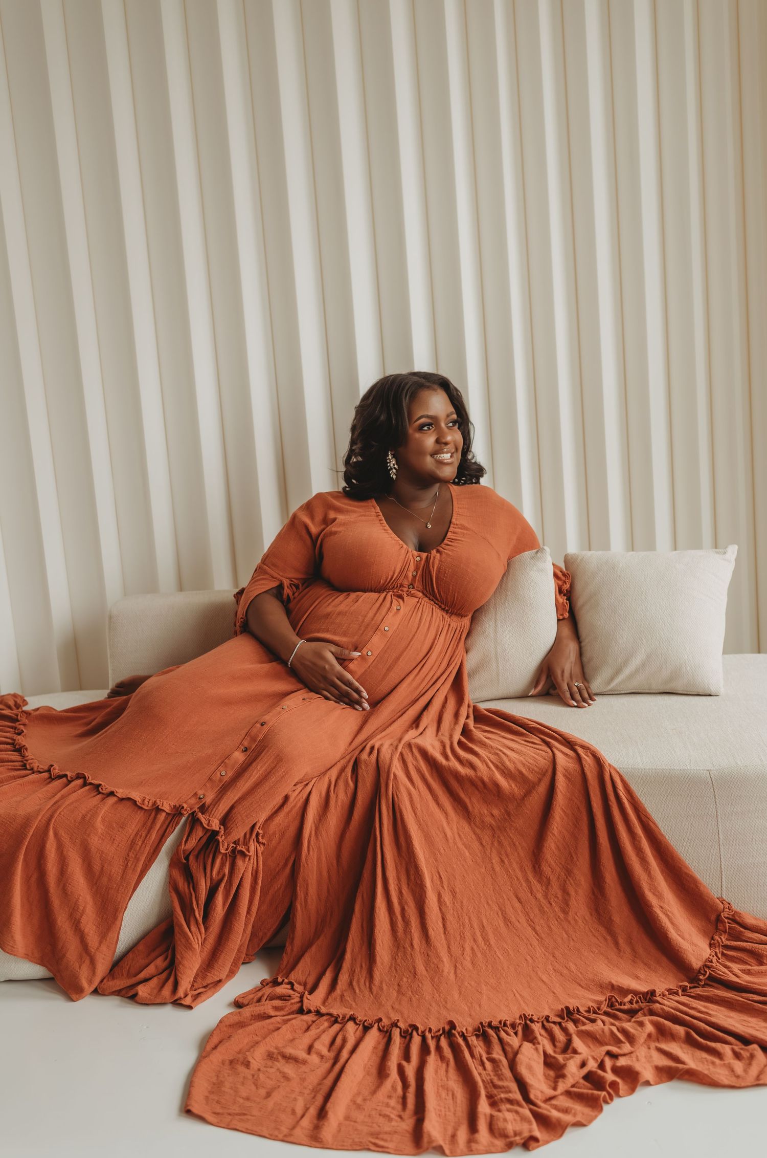 Pregnant woman in off-shoulder black lace gown smiling on vintage velvet sofa with pampas decor.