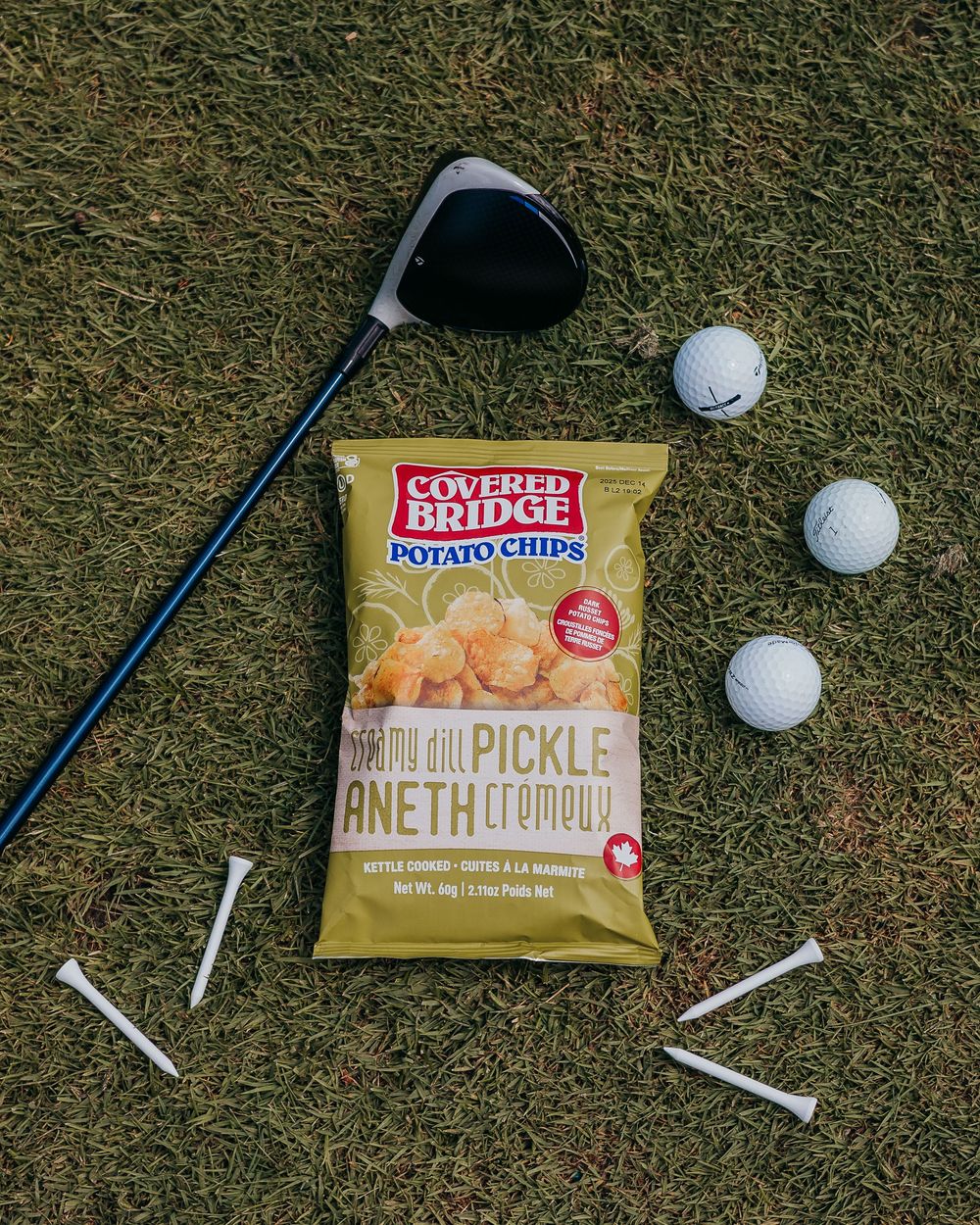 A golf club and scattered golf balls rest on green grass next to a bag of Covered Bridge Dill Pickle Chips.