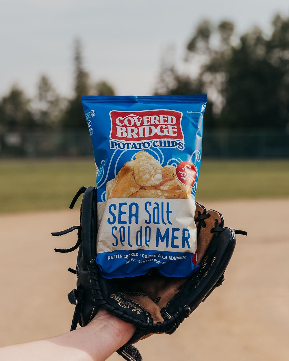Baseball glove in foreground with blue Covered Bridge potato chips bag against field background.