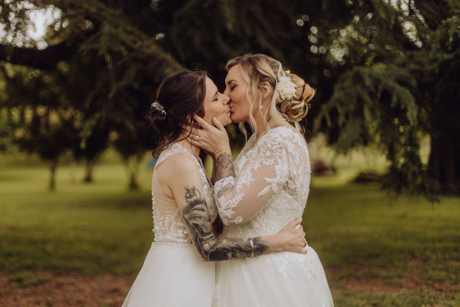 A romantic wedding moment between two brides in white lace dresses embracing outdoors under evergreen trees.