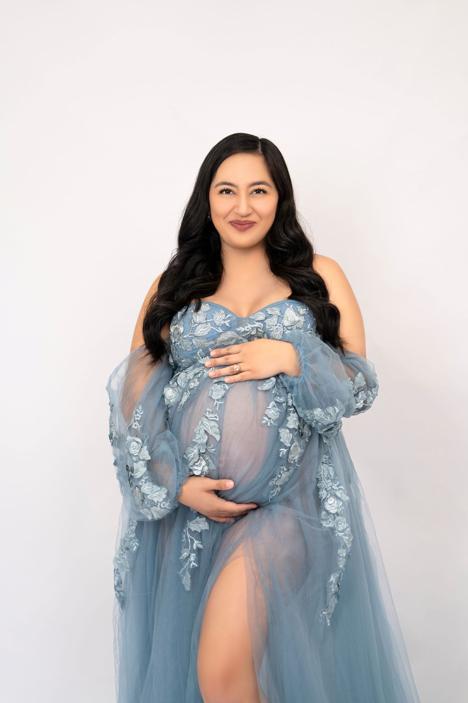 Maternity portrait in flowing blue floral gown against white backdrop.