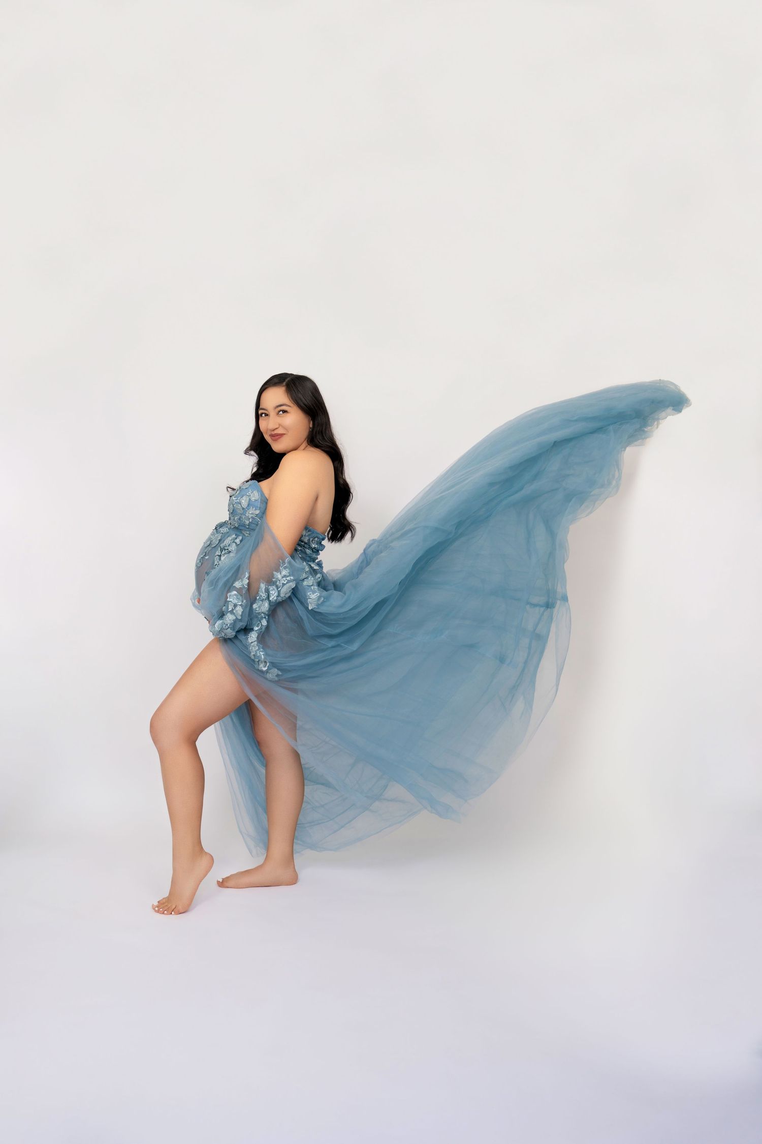 A figure in a flowing dusty blue tulle dress poses gracefully against a white backdrop.
