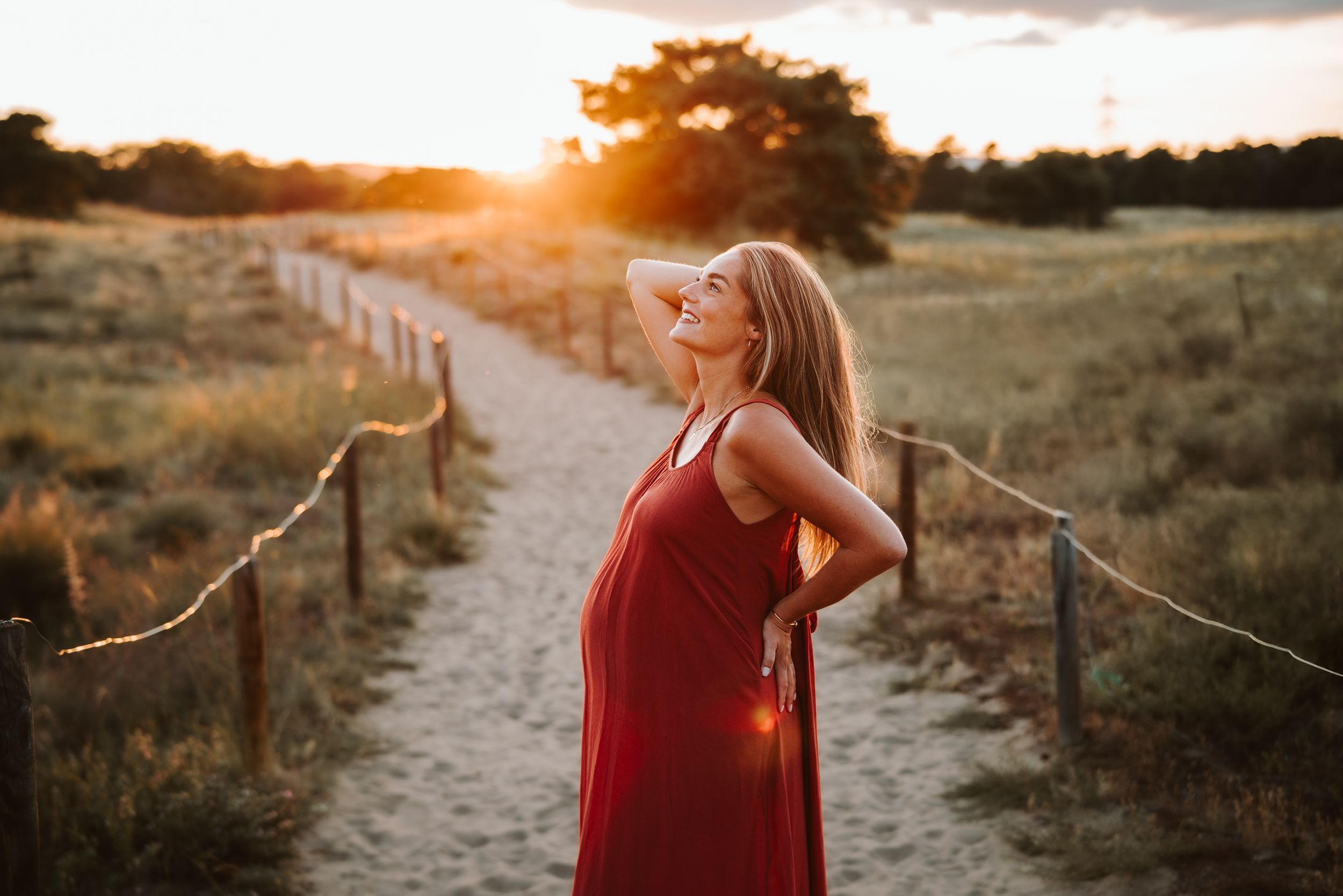 Maternity photography in red dress during golden hour in rustic outdoor setting.