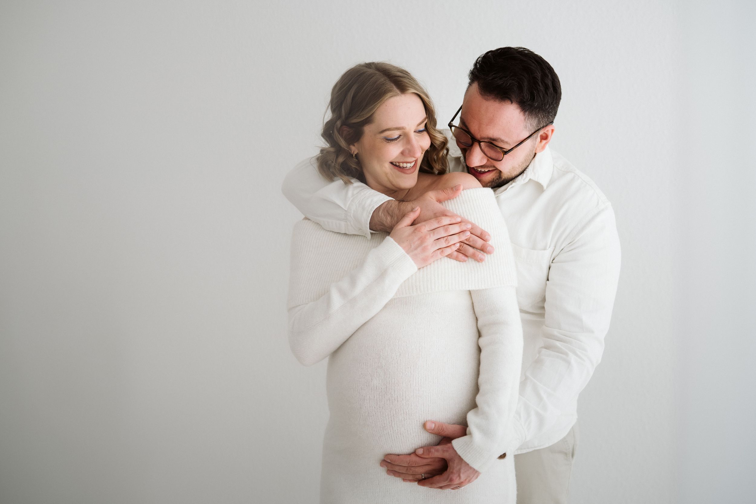 Joyful couple in white embracing during indoor maternity photoshoot.