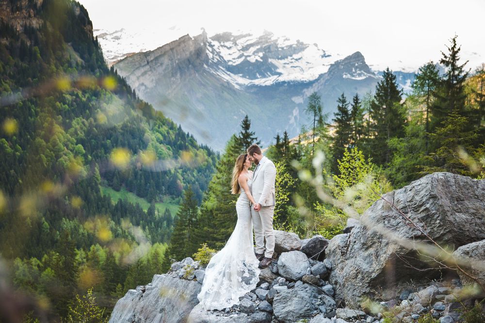 Wedding couple poses on mountain cliff with snow-capped peaks in background.